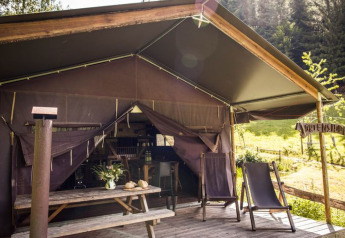 Tente safari avec terrasse ouverte, table en bois, chaises longues et vue sur la forêt verdoyante.