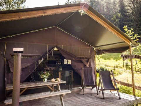 Safaritent met open veranda, houten picknicktafel, ligstoelen en uitzicht op bosrijke heuvels.