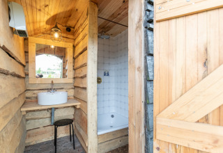 Salle de bain rustique d’une tente safari avec murs en bois, lavabo, miroir et douche privée chez le fermier.