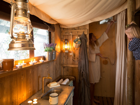 Vista interior de una tienda tipo safari con ducha privada, velas, faroles y dos mujeres en ambiente rústico.