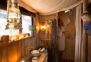 Vista interior de una tienda tipo safari con ducha privada, velas, faroles y dos mujeres en ambiente rústico.