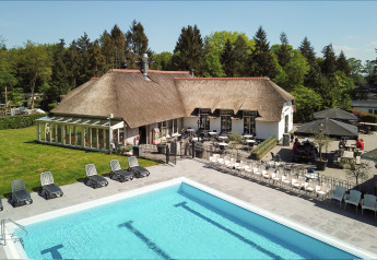 Piscina all'aperto con lettini di fronte a un alloggio glamping con tetto in paglia e terrazza, circondato da alberi.