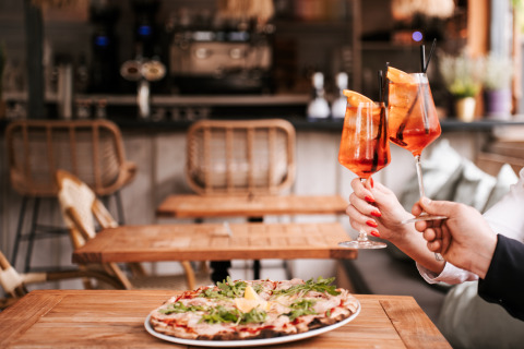 Dos personas brindan con cócteles sobre una pizza en una mesa de madera en un restaurante de glamping acogedor.