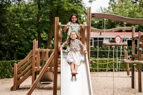 Dos niños jugando en un parque infantil de glamping, uno se desliza por el tobogán mientras el otro observa.