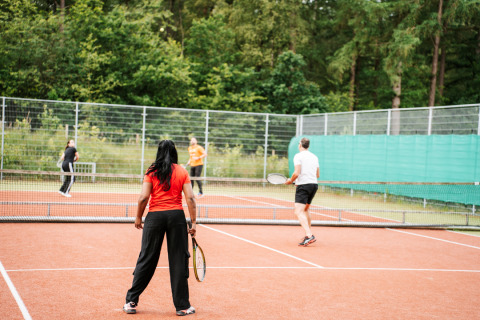 Vier Personen spielen Tennis auf einem Außenplatz inmitten von Bäumen bei einer Glamping-Unterkunft.