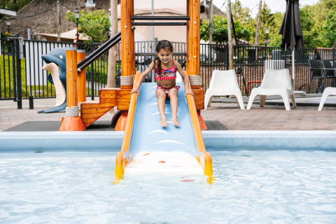 Young girl in swimsuit sliding down a water slide into a pool at a glamping site with outdoor seating.