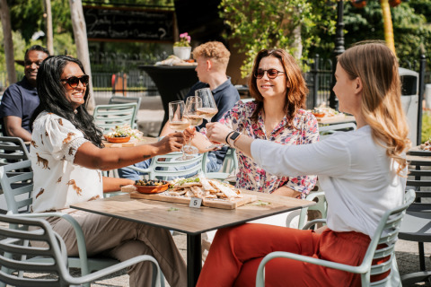 Tres mujeres brindan con copas de vino en una mesa exterior de glamping, rodeadas de comida y naturaleza.