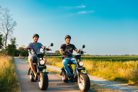 Dos hombres sonrientes pasean en scooters eléctricos por un camino junto a un alojamiento glamping en verano.