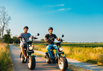 Due uomini sorridenti viaggiano su scooter elettrici lungo una strada vicino a un glamping sotto il sole estivo.