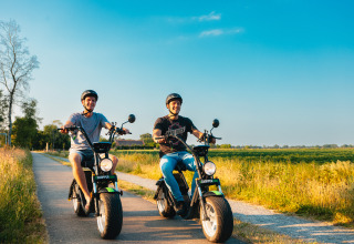 Deux hommes souriants roulent en scooter électrique sur un chemin de campagne près d’un site de glamping en été.