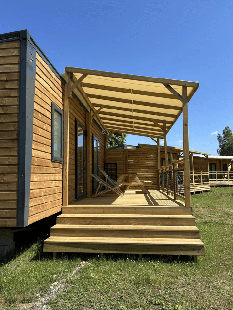 Holzhütte mit Veranda und Stühlen auf Camping Seasonova Les Marguerites in Frankreich bei Sonnenschein.