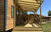 Holzhütte mit Veranda und Stühlen auf Camping Seasonova Les Marguerites in Frankreich bei Sonnenschein.
