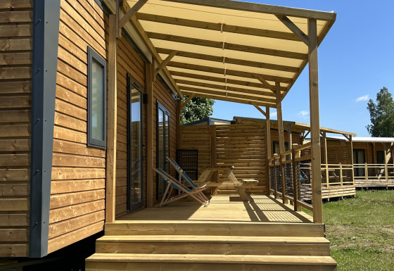 Holzhütte mit Veranda und Stühlen auf Camping Seasonova Les Marguerites in Frankreich bei Sonnenschein.
