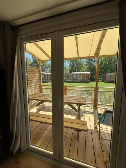Blick durch das Fenster einer Hütte im Camping Seasonova Les Marguerites, Frankreich, auf eine Holzterrasse.