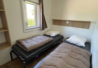 Innenansicht einer Hütte mit zwei Einzelbetten, braunen Decken, einem Fenster und Holzboden im Camping Seasonova Les Marguerites.