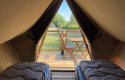 Inside a safari tent with two single beds, looking out onto a wooden deck and lush greenery outside.