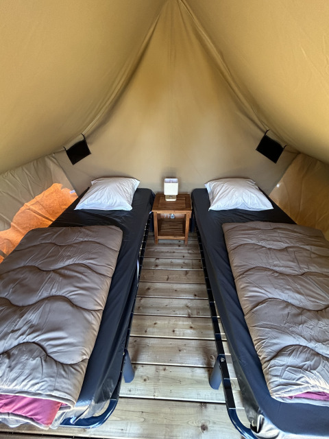 Vista interna della tenda safari Nomads Bivoac con due letti singoli, comodino e pavimento in legno al Camping Seasonova Les Marguerites in Francia.