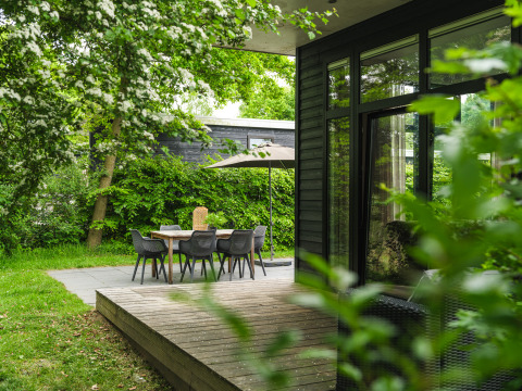 Zona de comedor al aire libre con mesa, sillas y sombrilla junto a una cabaña moderna rodeada de vegetación.