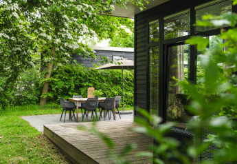 Udendørs spiseområde med bord, stole og parasol ved en moderne hytte, omgivet af frodig grøn bevoksning.