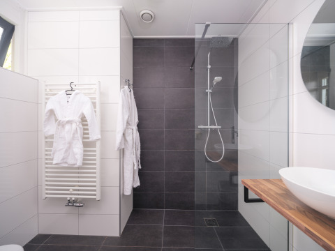 Moderne lodgebadkamer met glazen douche, houten werkblad, waskom en twee witte badjassen aan de muur.
