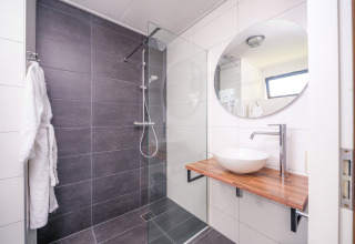 Bagno moderno in un lodge con grande doccia grigia, piano in legno, lavandino a conca e specchio tondo.