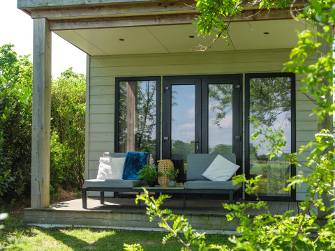 Knusse veranda bij een lodge met moderne zitbank, zachte kussens, planten en groen omringd.