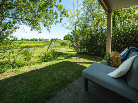 Blick von einer Lodge-Terrasse mit Sofa auf grüne Wiese, Bäume und einem Spielplatz am Wasser.