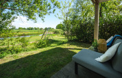 View from a lodge patio featuring a sofa, green garden scenery, and a swing set by a peaceful lake.