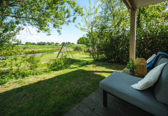 Vista dalla veranda di un lodge con divano, verde, alberi e altalena vicino a un laghetto tranquillo.