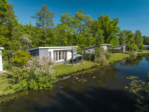 Lodge Mistique en Holiday Park De Lochemse Berg, Países Bajos, junto a un lago rodeado de naturaleza verde.