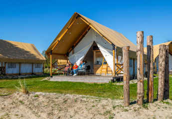 Gezin ontspant op de veranda van een Glampingtent Beachfront Luxe met helderblauwe lucht erboven.