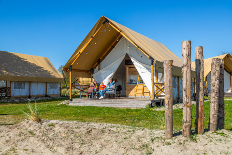 Familia relajándose frente a una Glampingtent Beachfront Luxe bajo un cielo azul brillante disfrutando del camping.