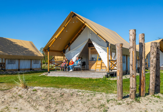 Familie sidder foran en luksuriøs Glampingtent Beachfront Luxe under blå himmel og nyder campinglivet.