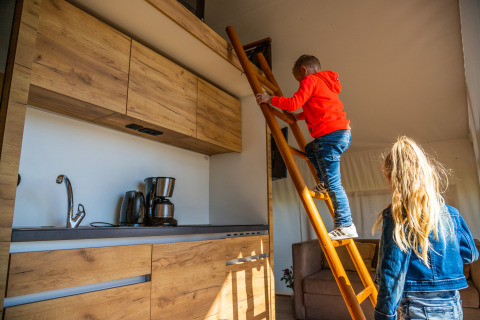 Two children exploring a loft area with a ladder in a modern kitchen of a beachfront glamping tent in Netherlands.