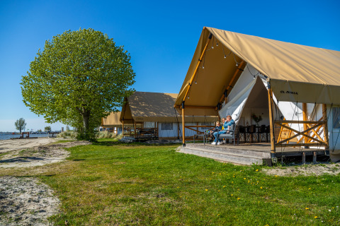 Tenda safari fronte mare allo Zuiderzee, Paesi Bassi, con persone rilassate in veranda sotto il cielo azzurro.