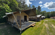 Safari-Zelte mit Sitzbereichen auf Holzterrassen stehen auf einer Wiese am Waldrand unter blauem Himmel.