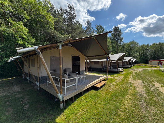 Safari-Zelte mit Sitzbereichen auf Holzterrassen stehen auf einer Wiese am Waldrand unter blauem Himmel.
