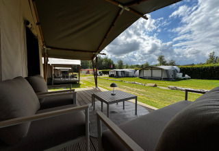 Vue depuis la terrasse d'une tente safari avec fauteuils et table, entourée de verdure et d'autres tentes.