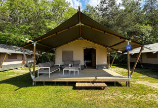Tienda safari en un camping con porche cubierto, asientos exteriores y entorno natural verde.