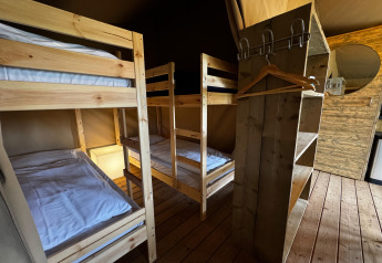 Interior de una tienda safari con dos literas de madera, estanterías, perchas y un espejo redondo.