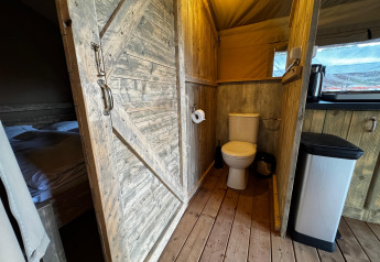 Vista interior de una tienda safari en Maasduinen, Países Bajos, con inodoro, cama y madera rústica.