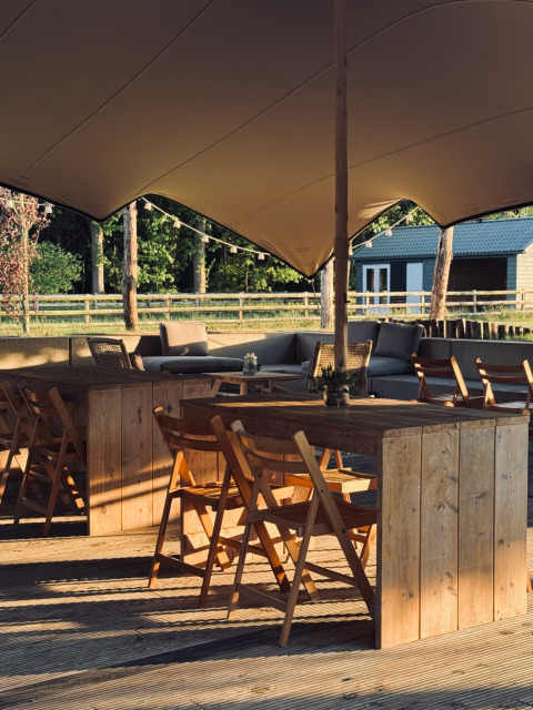 Outdoor seating area with wooden tables and chairs under canopy at Thuysgrond holiday park, North-Brabant, Netherlands.