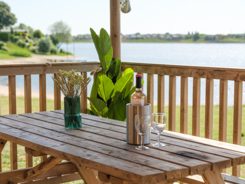 Holztisch auf einer Terrasse mit Weinflasche, Gläsern und Vase, Blick auf einen See in den Niederlanden.