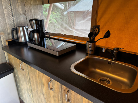 Plan de travail dans une tente safari avec bouilloire, cafetière, plaque, ustensiles et évier métallique.