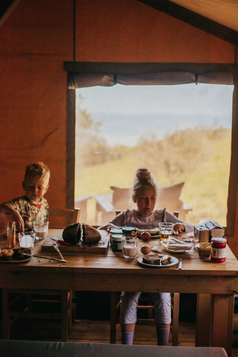 Twee kinderen ontbijten aan een houten tafel in een safaritent, met brood, eieren en glazen drinken.