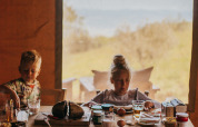 Twee kinderen ontbijten aan een houten tafel in een safaritent, met brood, eieren en glazen drinken.