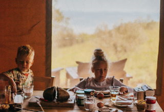 Two children sit at a wooden table inside a safari tent, enjoying breakfast with bread and drinks.