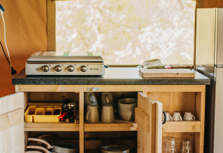 Camp kitchen at Safari Tent Garden, Camping Lucherino, Italy, with open cabinets showing pots, dishes, and mugs.