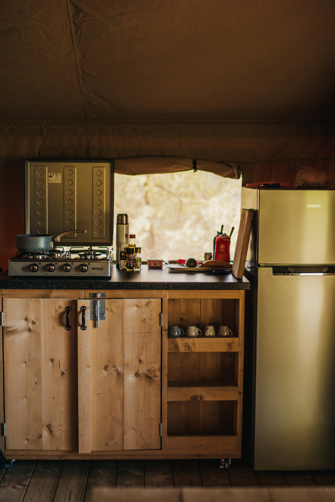 Angolo cucina in tenda safari con mobile in legno, fornello, tazze e frigo, Camping Lucherino, Italia.