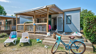 Knusse lodge met houten terras, ligstoelen, fiets en tuin op een zonnige dag.